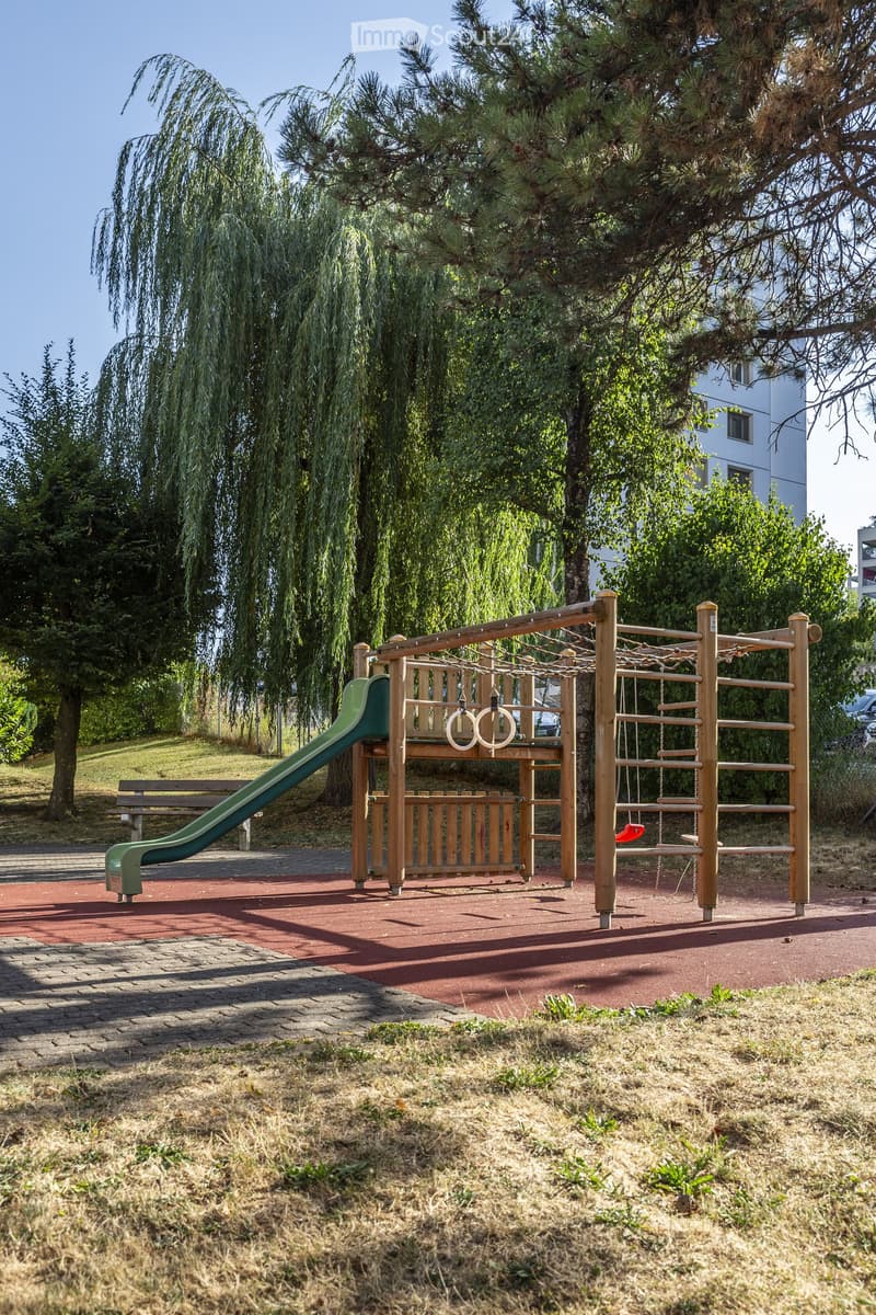 Playground with wooden climbing frame, green slide, swings, and a bench, surrounded by trees and grass.