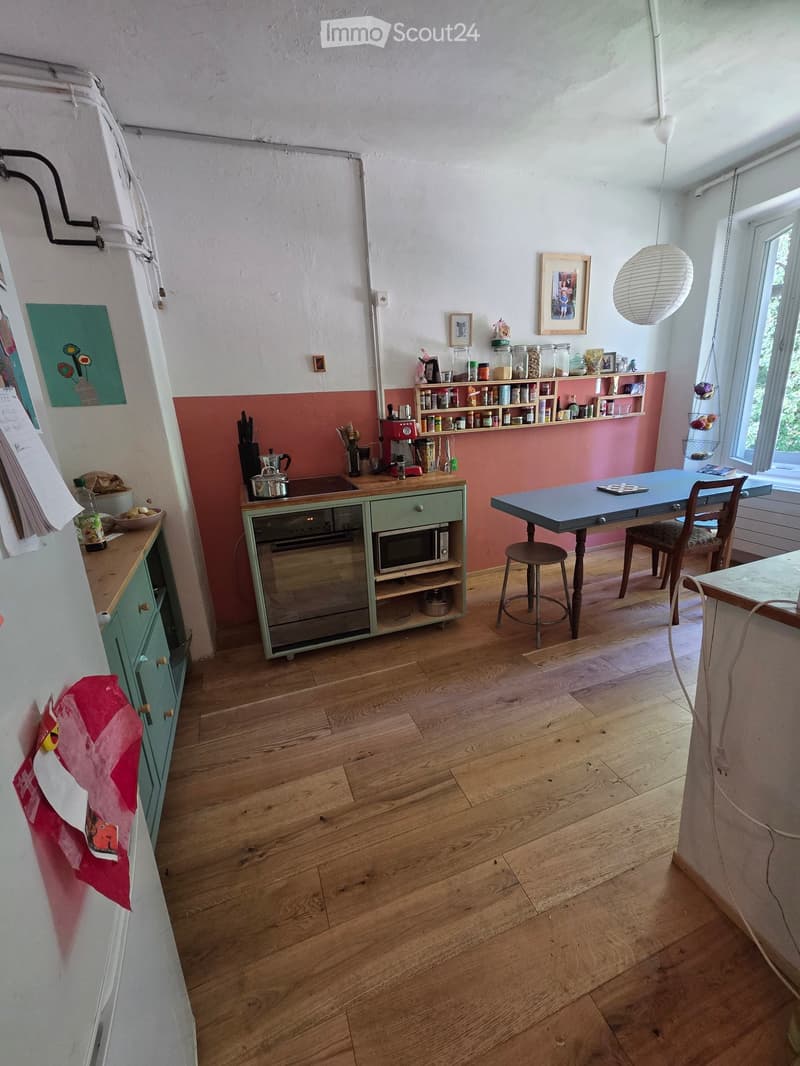 Bright kitchen with wooden flooring, light green cabinets, and a blue dining table with chairs. Shelves hold jars and kitchen items, and a pendant light hangs from the ceiling.