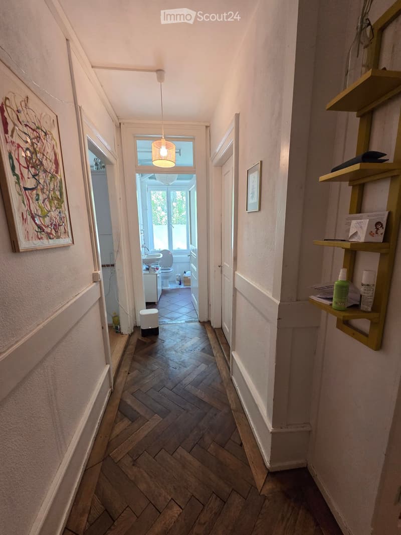 Narrow hallway with wooden herringbone flooring, white walls, and a pendant light fixture. A small shelf on the right holds cleaning supplies, and a painting hangs on the left wall. A bathroom is visible at the end of the hallway.