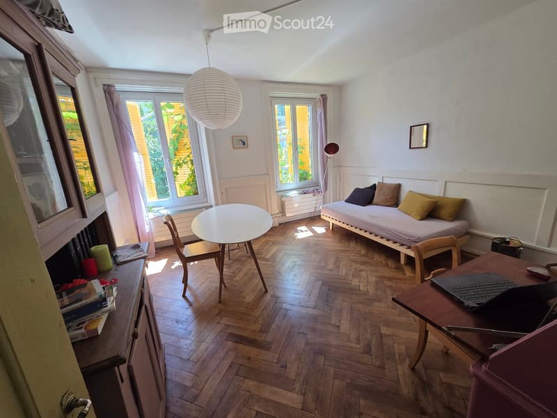 A cozy, well-lit room with wooden flooring, a daybed, a round table, a laptop on a desk, and a hanging spherical lamp.