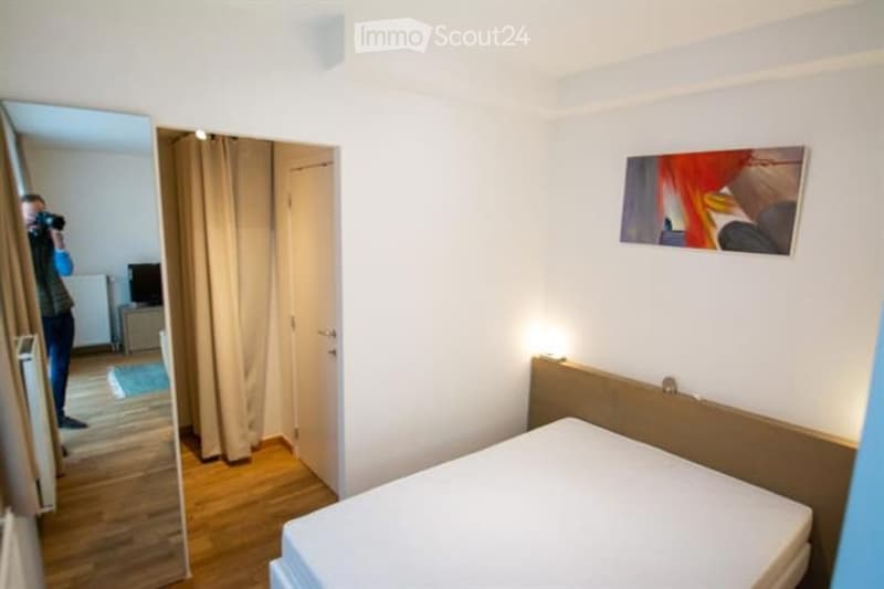 A modern, well-lit bedroom with a wooden bed frame, a large mirror, a painting on the wall, and a curtained doorway leading to another room.