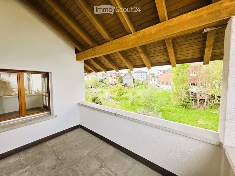 A balcony with a wooden ceiling, white walls, and a tiled floor. It has a window with wooden frames and a view of a grassy area with trees and houses.