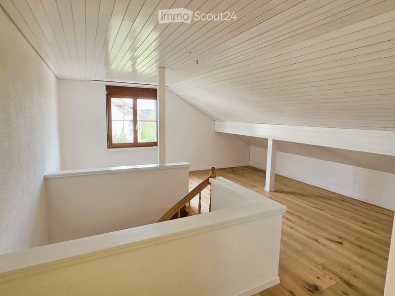 An empty attic room with wooden floors, white walls, and a wooden staircase. There is a window with wooden frames.