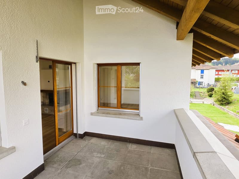 A balcony with a tiled floor, white walls, and a wooden ceiling. There is a glass door leading to the interior and a window with wooden frames.