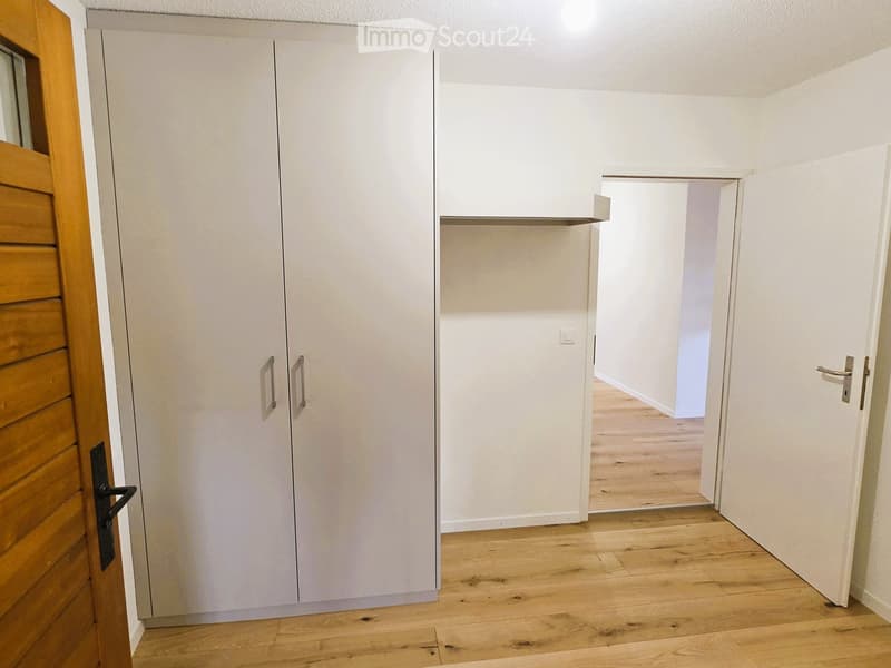An empty room with white walls, wooden floor, and a large white wardrobe with two doors and a shelf above it. There is a wooden door on the left and an open door on the right leading to another room.