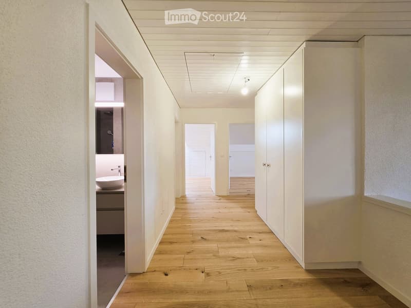 Empty corridor with wooden floor, white walls, and ceiling lights. A bathroom is visible through an open door, featuring a sink and faucet.