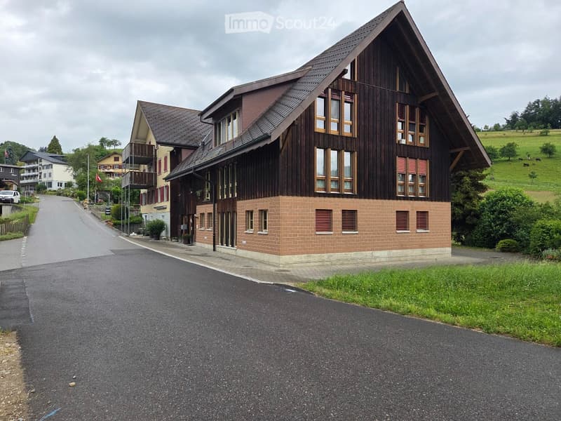 A two-story residential building with a wooden facade and brick base, featuring multiple windows and a sloped roof. The building is situated on a paved road with surrounding greenery and neighboring houses visible in the background.