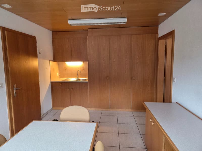 A small, well-lit room with a wooden cabinet, a sink, a chair, and a table. The room features tiled flooring and a wooden ceiling with integrated lighting.