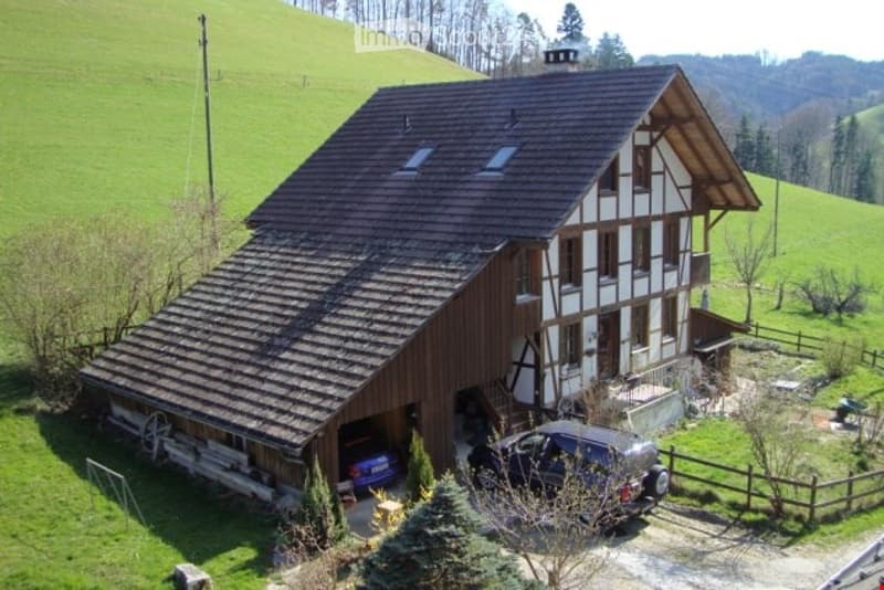 A two-story house with a wooden exterior and dark roof, situated on a hillside with a garage and parking area.