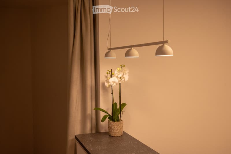 A modern, minimalist room with a sleek black table, a white orchid in a woven pot, and a set of three pendant lights suspended from the ceiling.