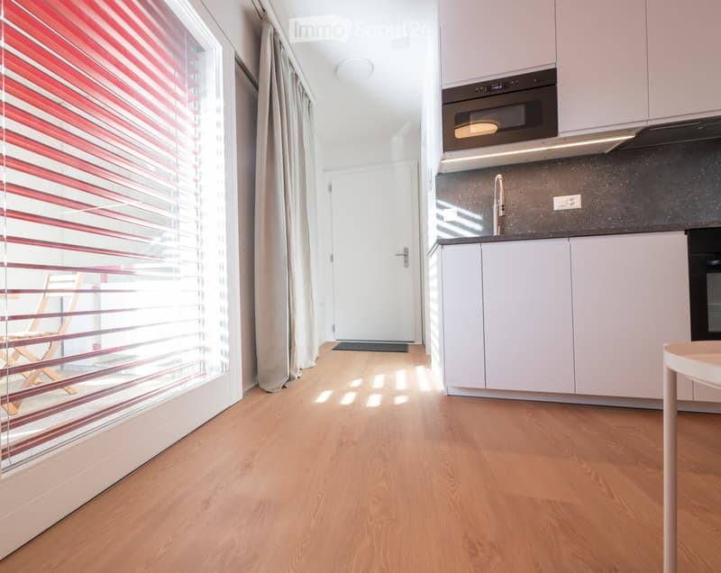 Modern kitchen with white cabinets, stainless steel appliances, and a wooden floor. A small stool and a chair are visible near the window.