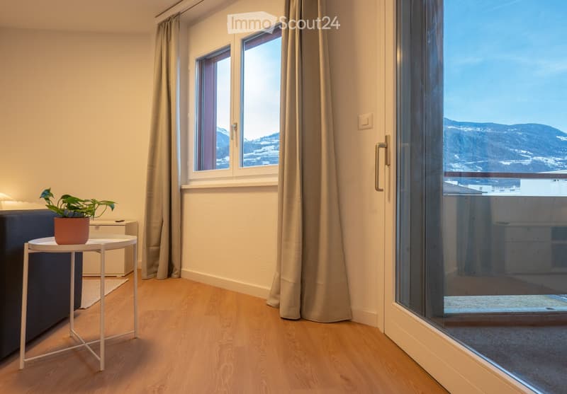 Bright, modern room with wooden flooring, large windows, and a balcony offering a scenic mountain view. Features a sofa, small round table with a potted plant, and light-colored curtains.