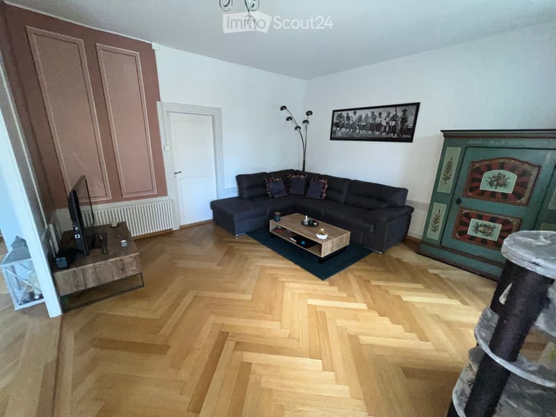 Modern living room with wooden herringbone flooring, dark sectional sofa, wooden coffee table, flat-screen TV on a wooden stand, and a decorative cat tree.