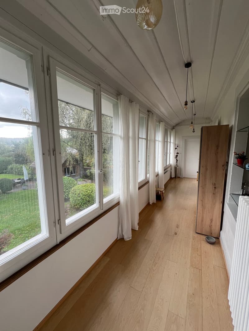 Bright hallway with wooden flooring, large windows with white curtains, and a view of a green garden outside.