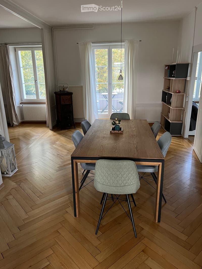 Dining room with wooden herringbone flooring, large windows with white curtains, a modern dining table with six chairs, a shelf unit, and a lantern on the floor.