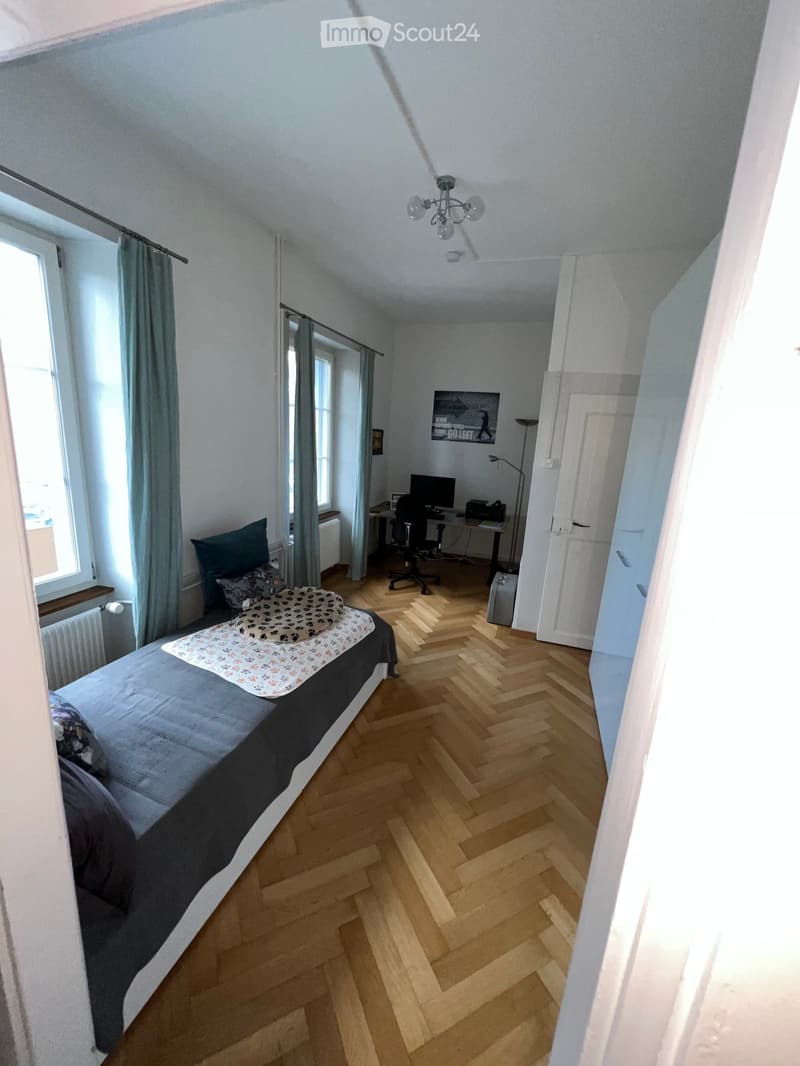 Bedroom with wooden herringbone flooring, a single bed with pillows and a blanket, a desk with a computer and chair, and large windows with curtains.