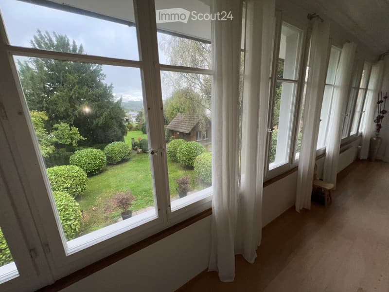 Bright hallway with large windows and white curtains, leading to a view of a well-maintained garden with shrubs and a small shed.
