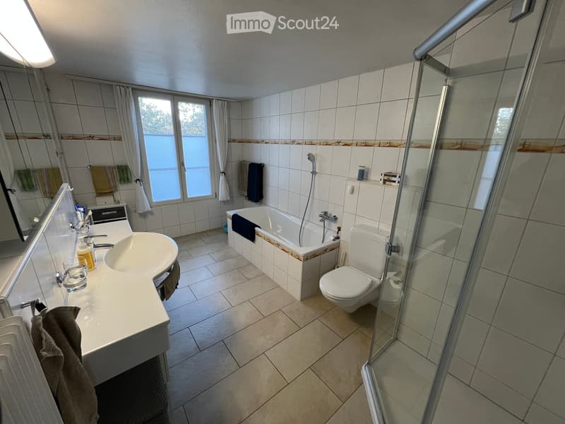 Modern bathroom with a white sink, bathtub, toilet, and glass-enclosed shower. The room features tiled walls and floor, a large window with curtains, and various towels and toiletries.