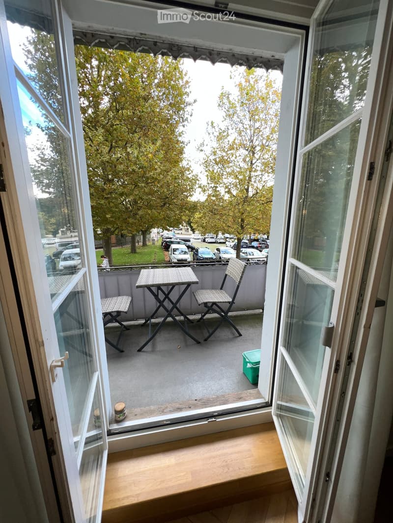 Balcony with a small table and two chairs, overlooking a street with parked cars and trees.