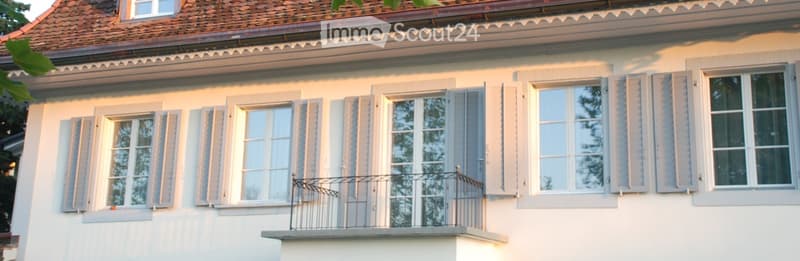 Two-story building with white exterior, multiple windows with shutters, and a small balcony with metal railing.