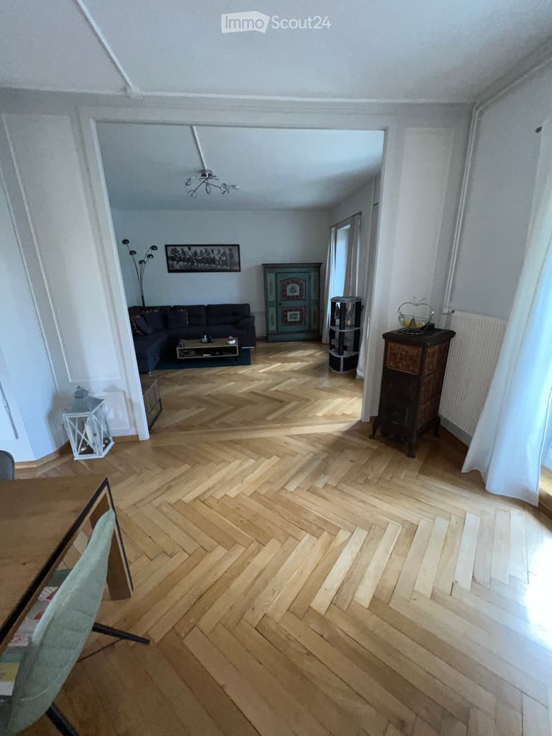 Living room with herringbone wooden flooring, white walls, a dark blue sofa, a coffee table, a small table with a chair, a cabinet, a shelf, a picture frame, a floor lamp, and a radiator.