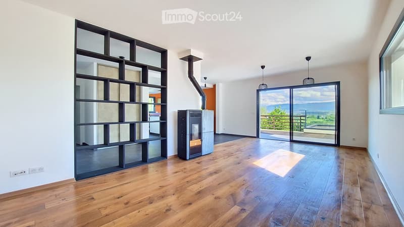 Empty room with wooden flooring, white walls, large glass doors leading to a balcony with scenic mountain view, modern shelving unit, and a wood-burning stove.