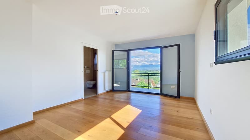 Empty room with wooden flooring, white walls, large sliding glass doors leading to a balcony, and an adjacent bathroom visible through an open doorway.