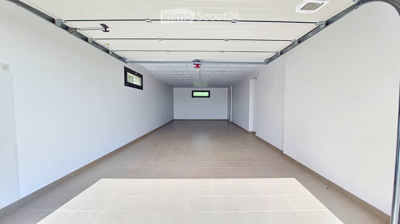 Empty corridor with white walls, tiled floor, and a partially open garage door at the end.