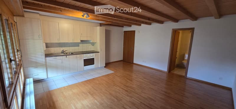 Bright, open room with wooden floors, white cabinets, a sink, and an oven in the kitchen area. A separate bathroom with a toilet is visible through an open doorway.