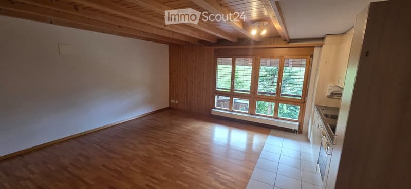 Empty room with wooden flooring, white tiled kitchen area, large windows with blinds, and exposed wooden ceiling beams.