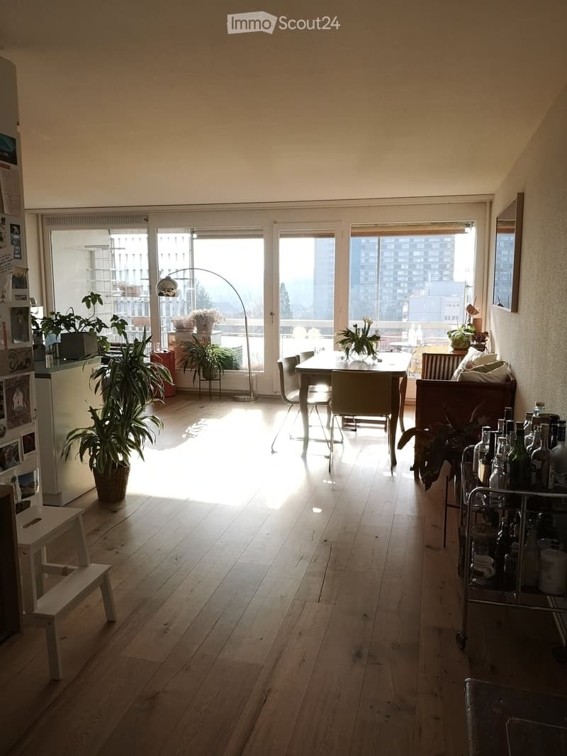 Bright living space with wooden flooring, large windows, a dining table with chairs, a sofa, a floor lamp, potted plants, and a wine rack.