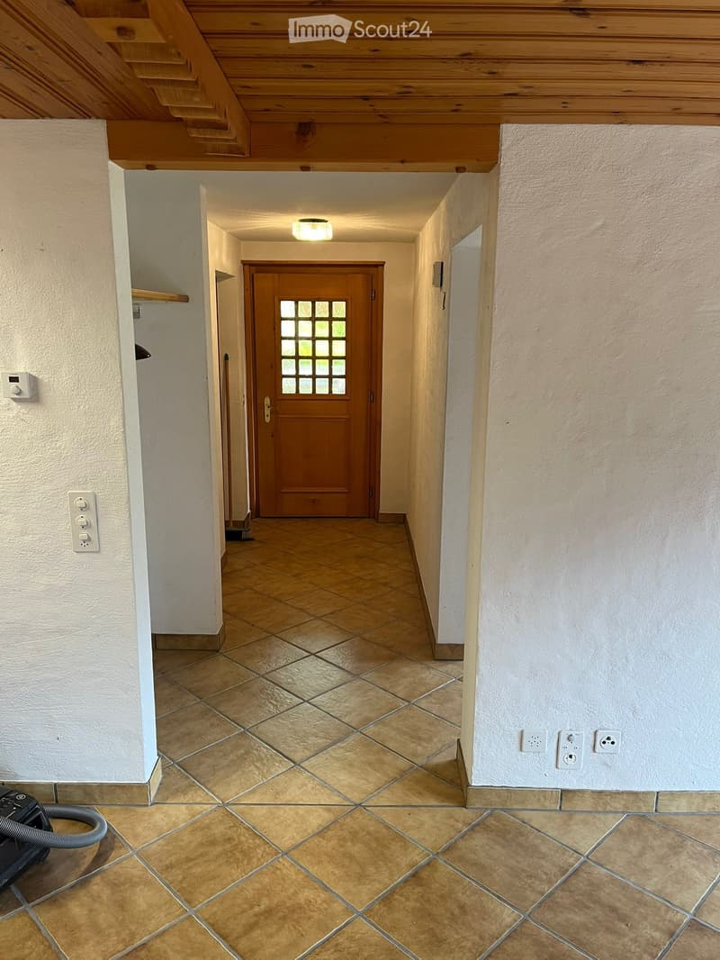 A clean, well-lit hallway with tiled flooring, white walls, and a wooden door at the end. Electrical outlets and a thermostat are visible on the walls.