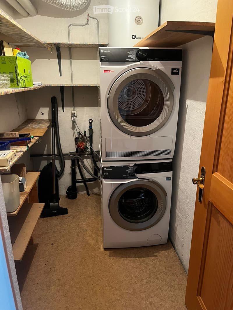 Compact laundry area with a stacked washing machine and dryer, shelves, and a vacuum cleaner.
