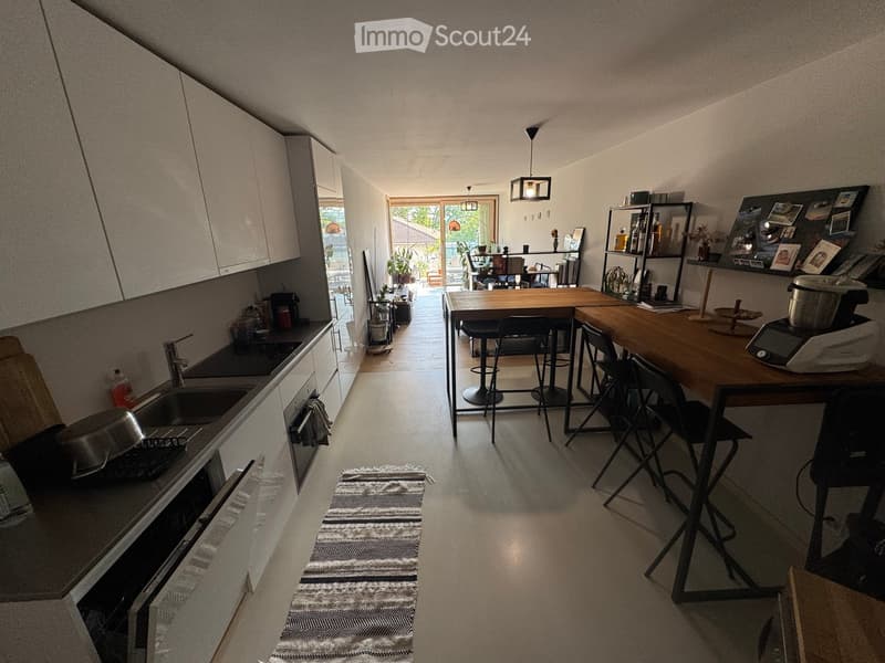 The image shows a modern, open-plan kitchen and dining area with white cabinets, a central wooden dining table, black chairs, and stools. The kitchen features a sink, oven, and various appliances. A rug is placed on the floor, and there is a view of a bal