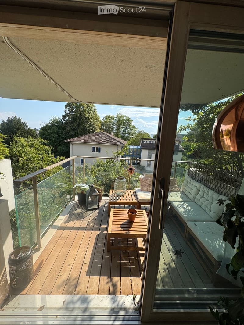 Balcony with wooden decking, glass railing, sofa, small table, chairs, lantern, and potted plants.