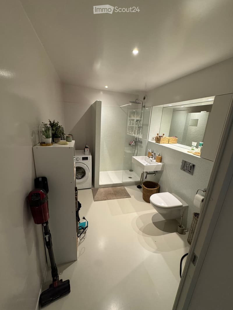Modern bathroom with white tiles, a toilet, sink, shower, washing machine, and decorative plants.