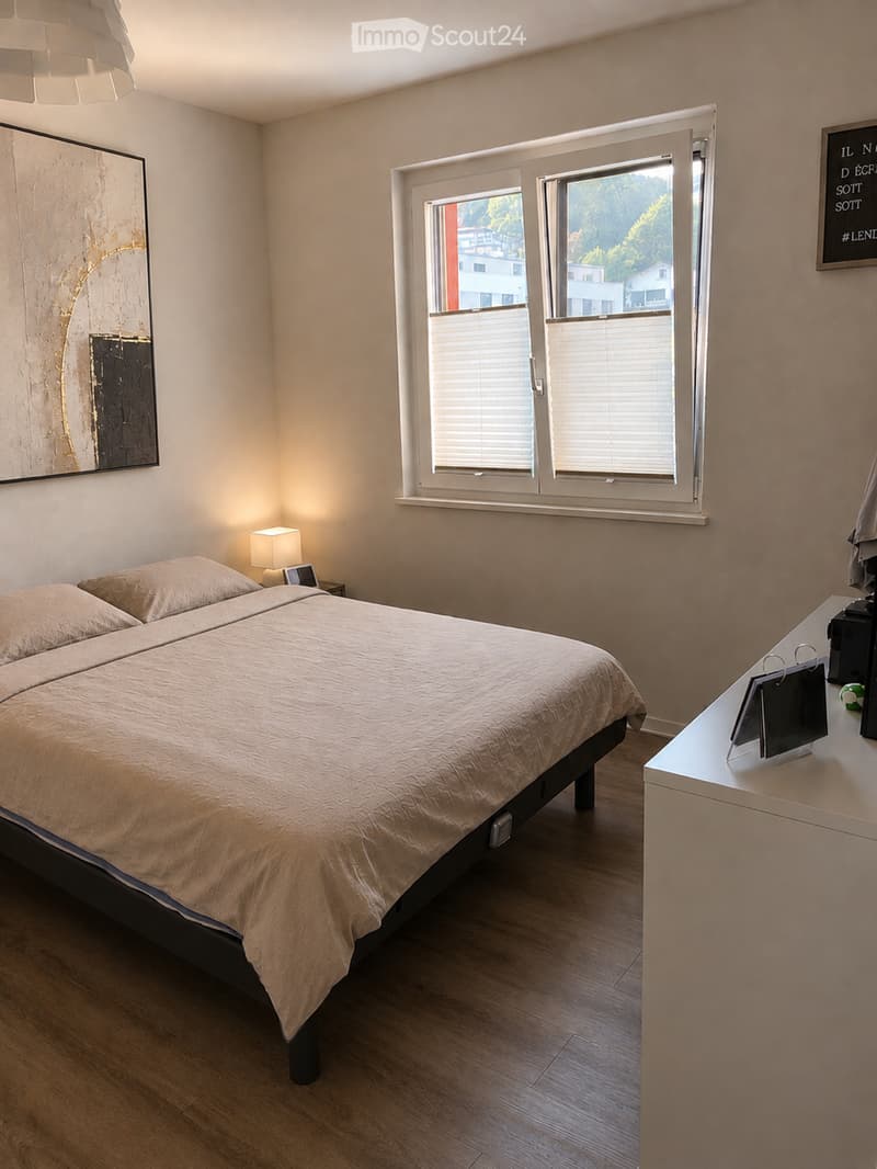 Cozy bedroom with a double bed, nightstand with lamp, large window with blinds, and a small table with a screen and other items.