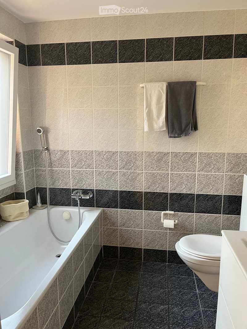 Modern bathroom with a bathtub, toilet, and towel rack. Features a mix of beige and black tiles, a shower hose, and a small basket on the windowsill.