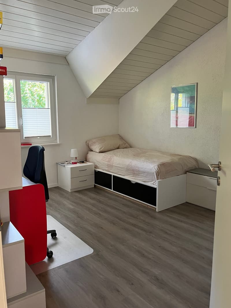 Cozy attic bedroom with a single bed, nightstand, lamp, chair, and mirror. The room features wooden flooring and slanted ceilings.