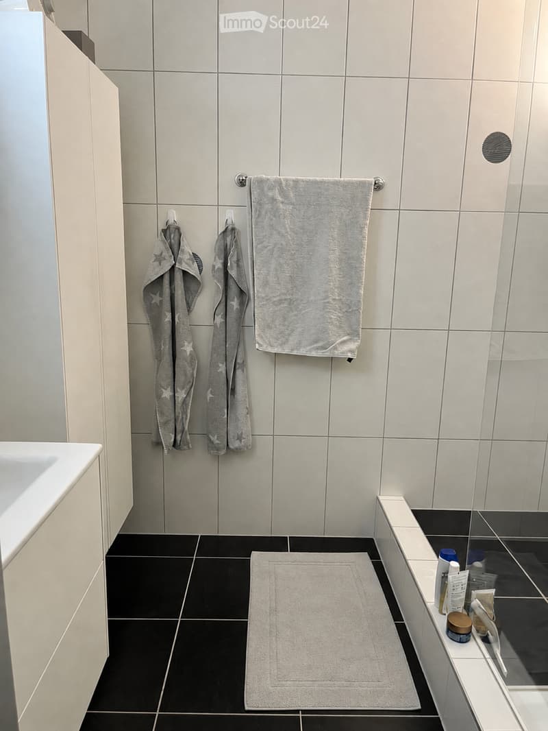 Modern bathroom with white tiled walls, black tiled floor, and a white cabinet. Features include two hanging towels, a bath mat, and toiletries on the edge of the tub.