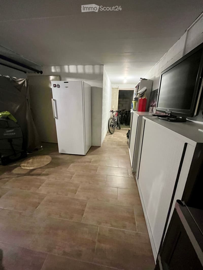 A narrow hallway with tiled flooring, white walls, and a refrigerator on the left side. A television is placed on a stand to the right, and bicycles are visible in the background.