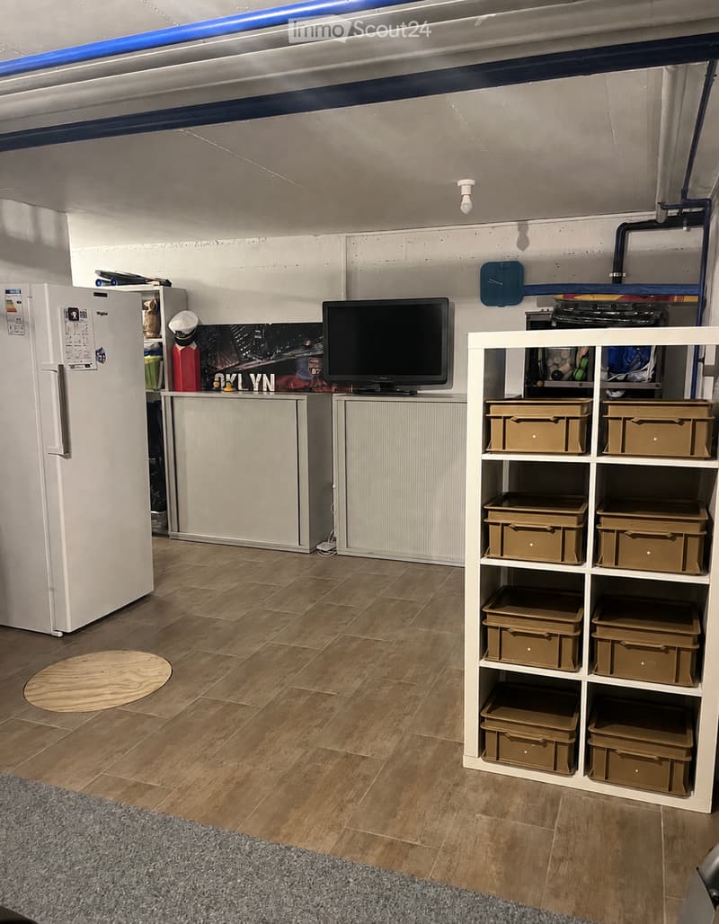 The room features a refrigerator, a television, a white shelving unit with storage boxes, and a wooden floor.