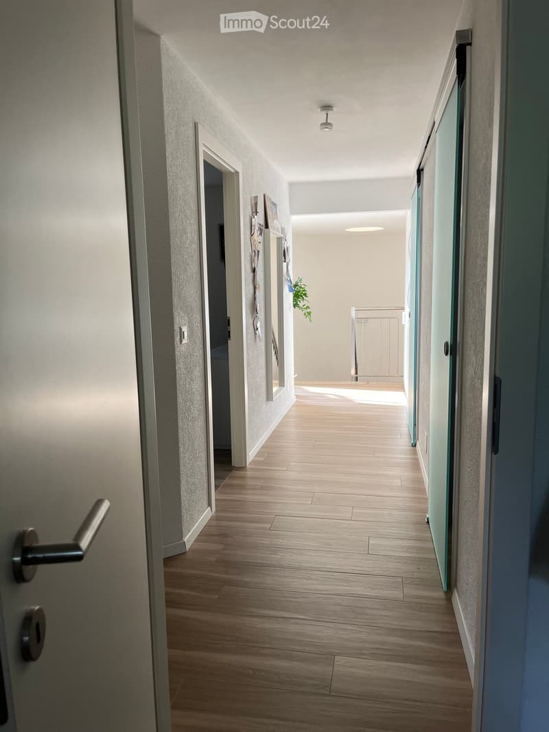 A modern hallway with wooden flooring, white walls, and recessed lighting. Features a glass door on the right and a partially visible room with a plant on the left.