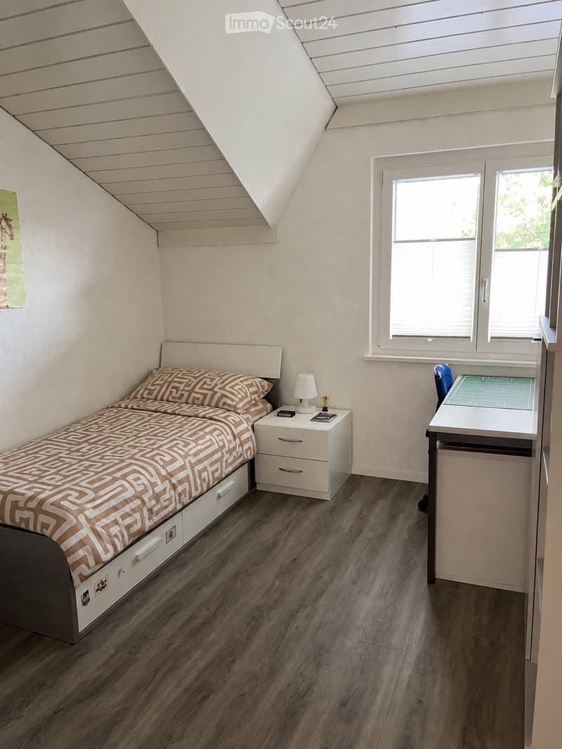 Cozy attic bedroom with wooden flooring, slanted ceiling, large window, bed with patterned bedding, nightstand with lamp, and desk with chair.