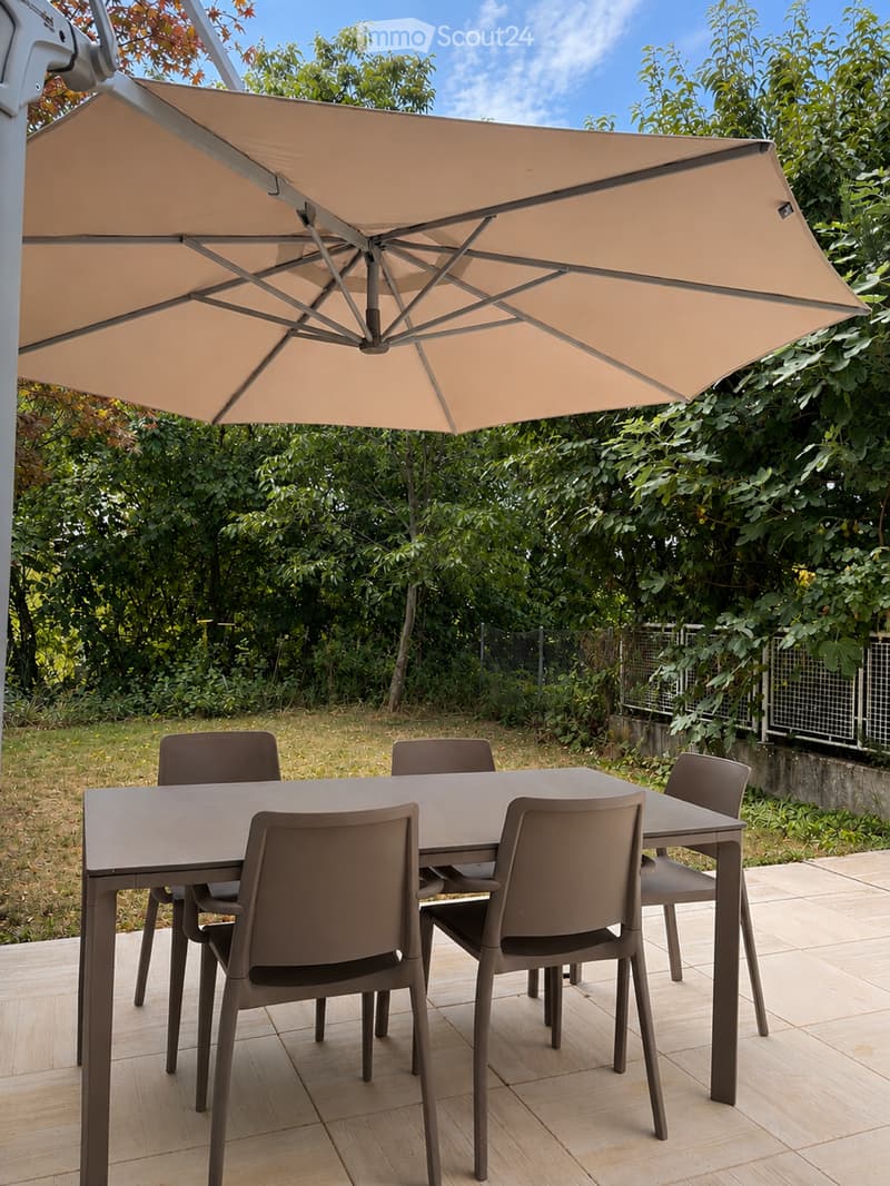 A modern outdoor dining set with a rectangular table and four matching chairs, placed on a wooden deck under a large beige umbrella, surrounded by lush greenery.