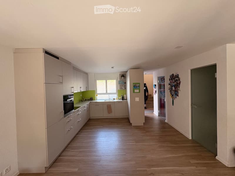 Bright hallway with wooden flooring leading to a modern kitchen area featuring white cabinets, a green backsplash, and stainless steel appliances.