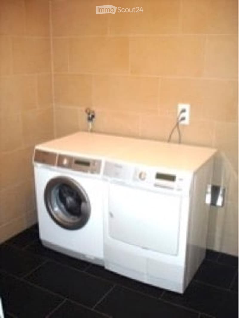 A compact bathroom with a white washing machine and dryer unit, beige tiled walls, and black tiled floor.