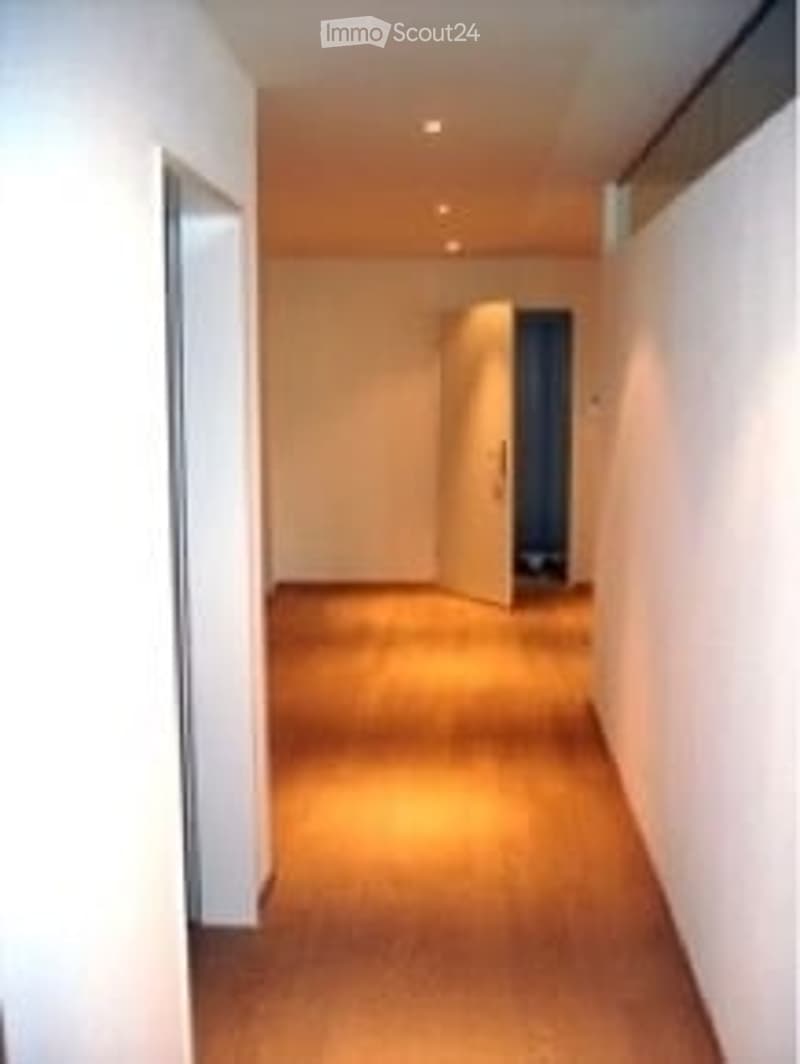 A well-lit hallway with wooden flooring, white walls, and recessed ceiling lights. The hallway leads to an open doorway at the end.