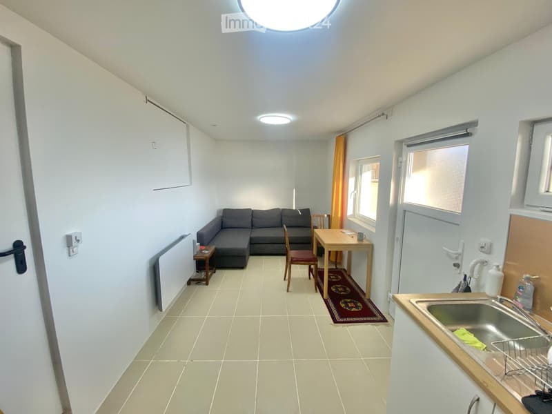 A compact, well-lit living space with a sofa, small table, chair, and a sink area. The room features tiled flooring, white walls, and large windows with blinds.