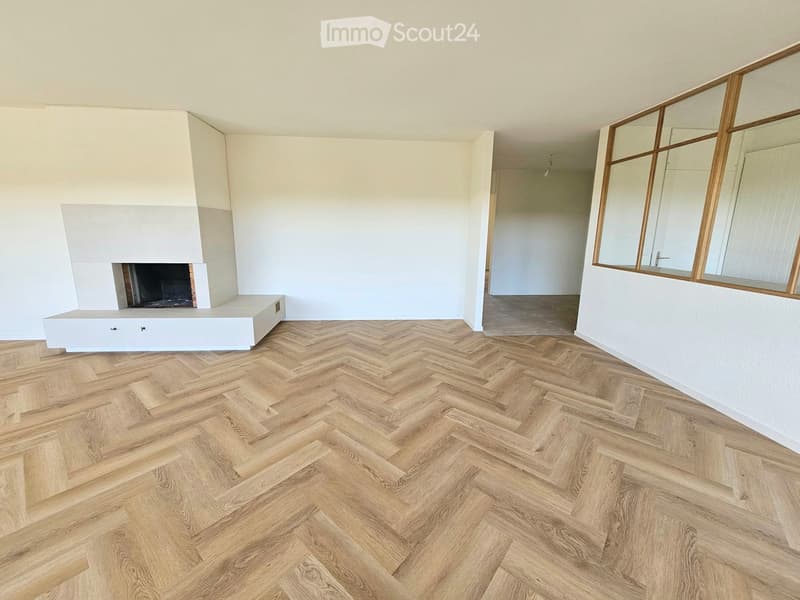 Empty room with herringbone wooden flooring, white walls, fireplace, and large mirror on the wall.